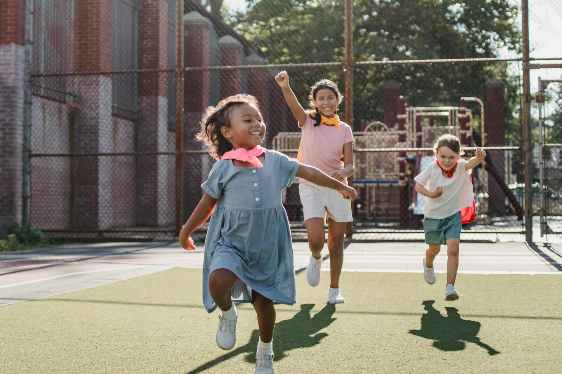 Three children wearing casual clothes run and play on an outdoor
sports court with a playground and fence in the background.