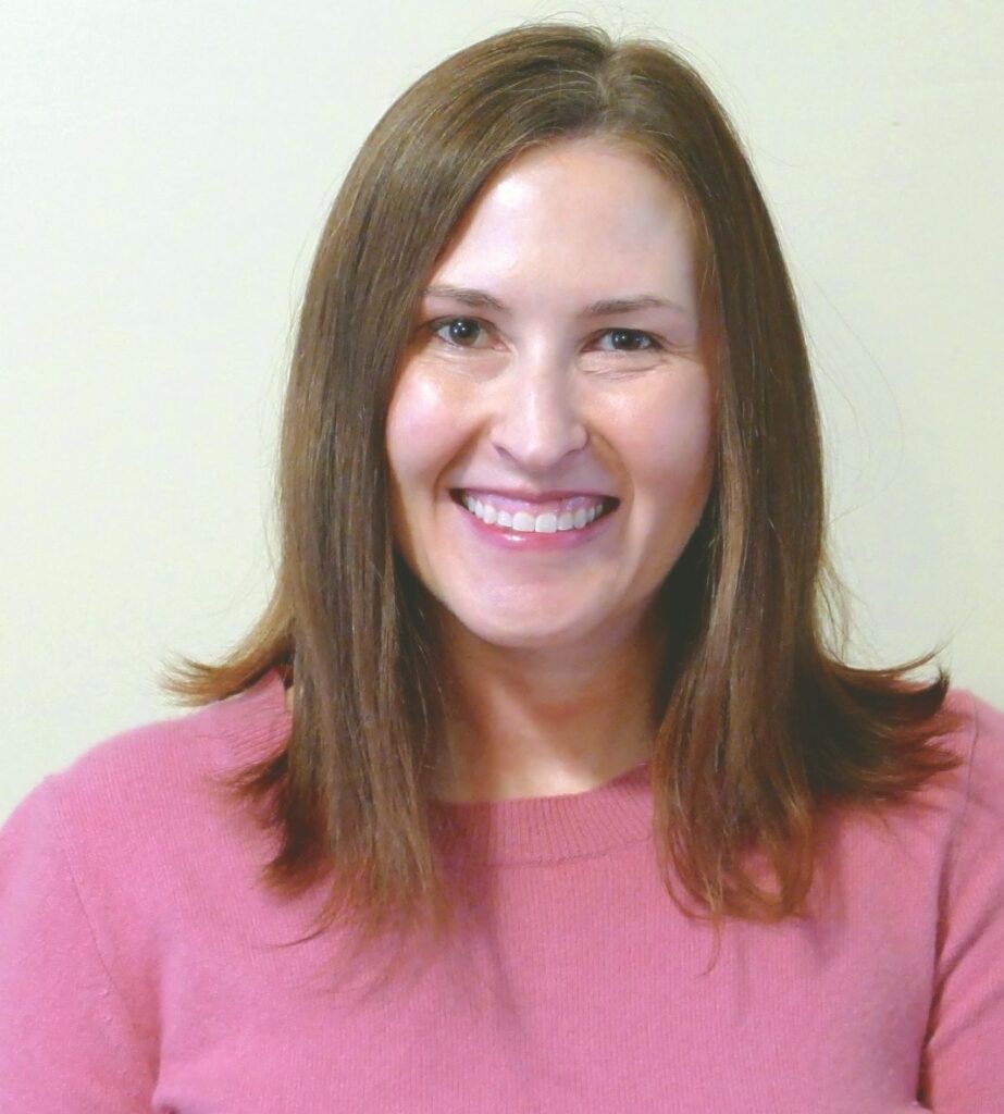 A woman with straight, shoulder-length brown hair and a pink sweater smiles warmly at the camera against a plain, light-colored background.