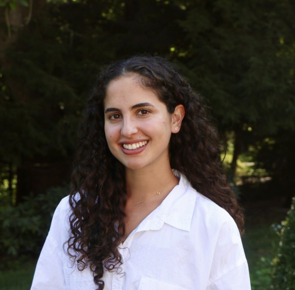 A young woman with long curly hair smiles at the camera. She is wearing a white shirt and standing outdoors with greenery and trees in the blurred background.