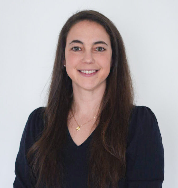 A woman with long brown hair wearing a black top with gold buttons on the sleeves stands against a plain white background, smiling at the camera with her hands folded in front of her.