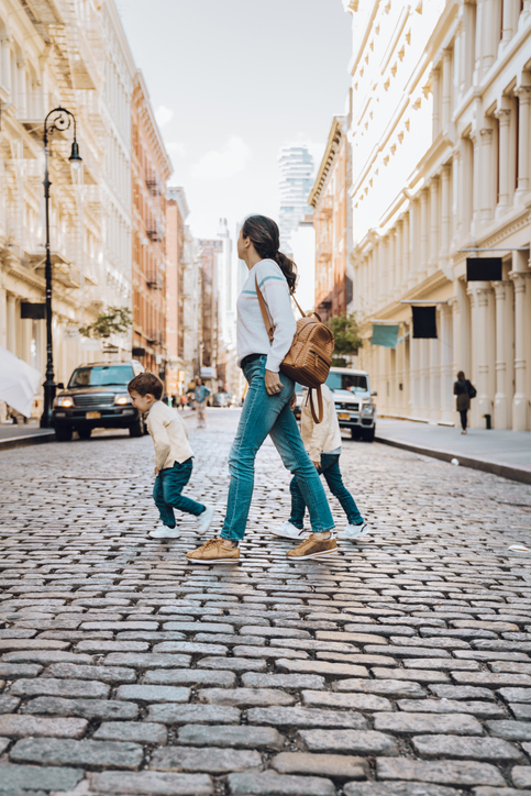 A woman with a backpack stands on a cobblestone street while two young children cross in front of her. The street is lined with tall buildings and cars are parked along the sides. The scene appears to be in a city.