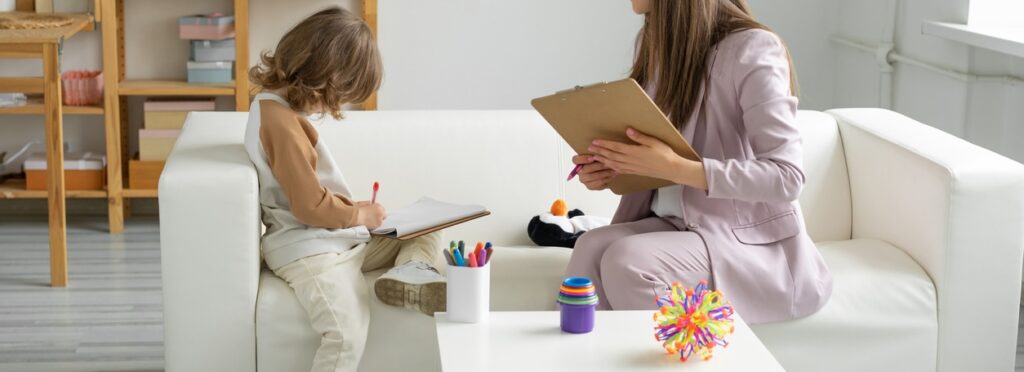 A child sits on a white sofa drawing on a clipboard, while an adult with a clipboard and pen sits nearby, possibly conducting a therapy or counseling session in a bright, organized room with toys and shelves.