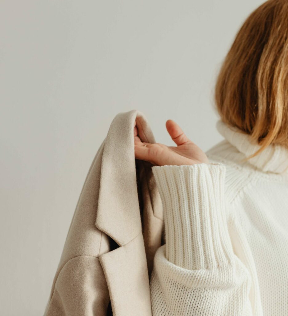 A person in a white knit sweater, seen from behind, holds a beige coat draped over their hand against a plain, light-colored background.