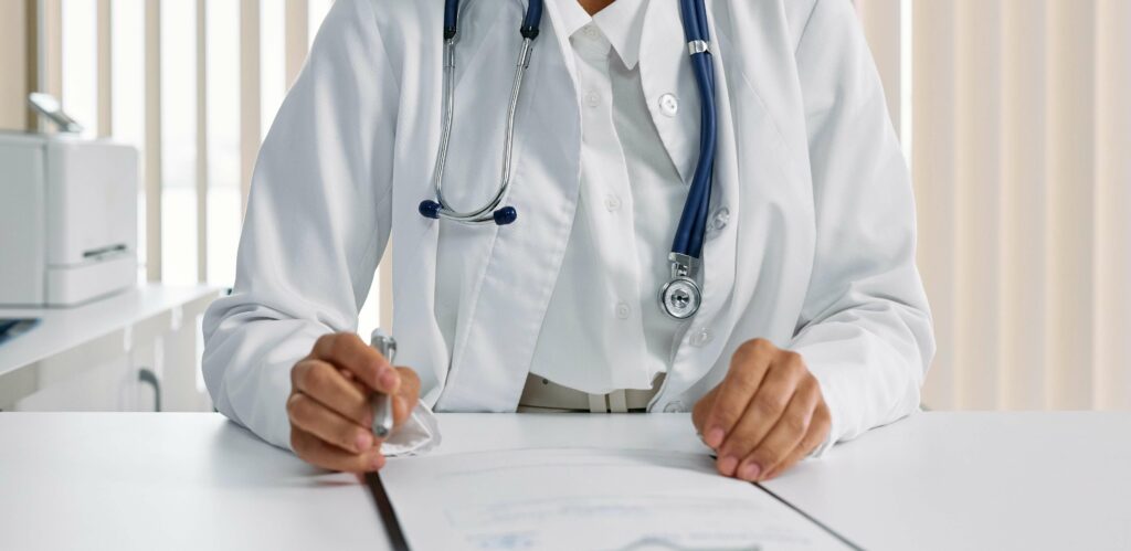 A doctor in a white coat with a stethoscope around their neck sits at a desk, holding a pen and reviewing paperwork. The face is not visible.