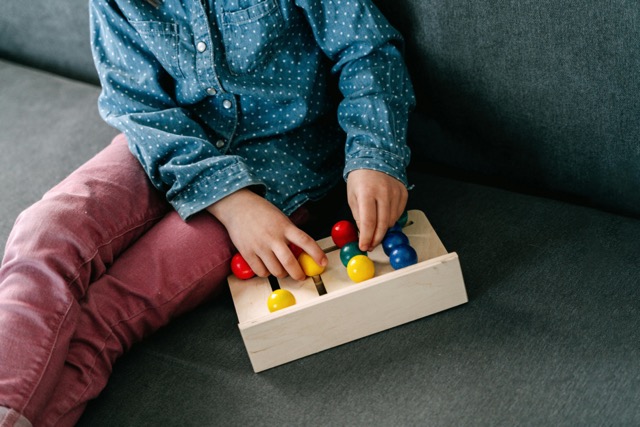 A child wearing a blue polka dot shirt and pink pants sits on a gray couch, playing with a wooden toy featuring colorful pegs and knobs.