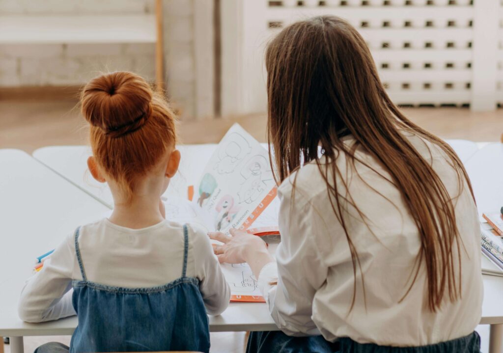 A young girl and an adult sit side by side at a table, looking at a colorful book together. Both have long hair, and the scene suggests a learning or tutoring activity in a bright room.