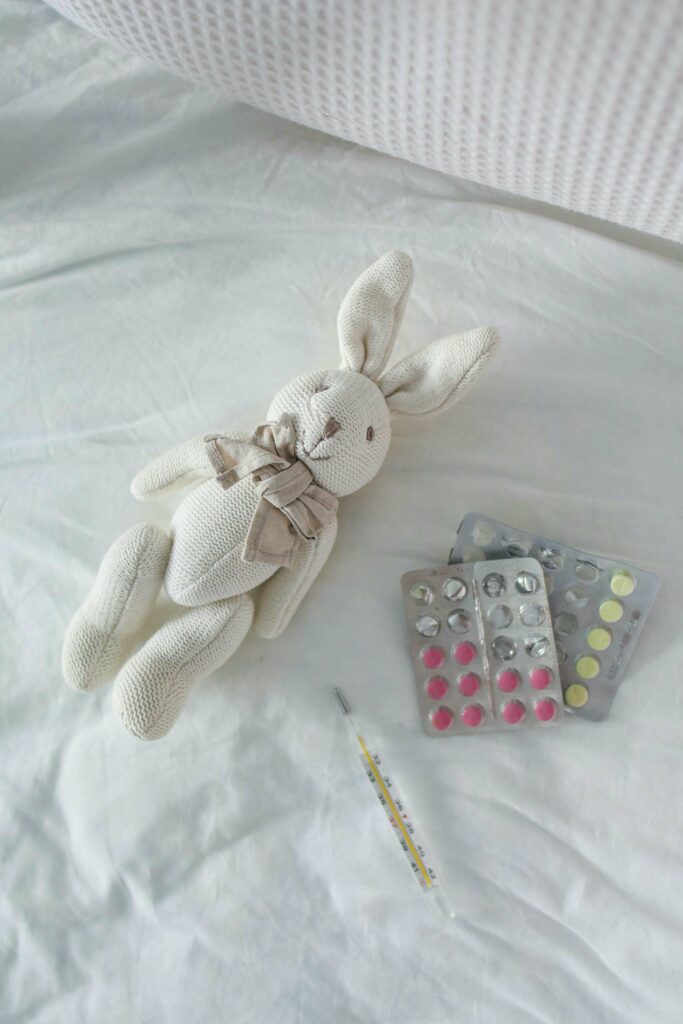 A stuffed white bunny with a bow lies on a bed next to a thermometer and several blister packs of colorful pills.
