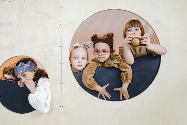 Four children playfully peer through round cutouts in a light wooden wall. Two are dressed as animals, one holds a toy telescope, and another hides behind a panel, suggesting imaginative play or a theater scene.