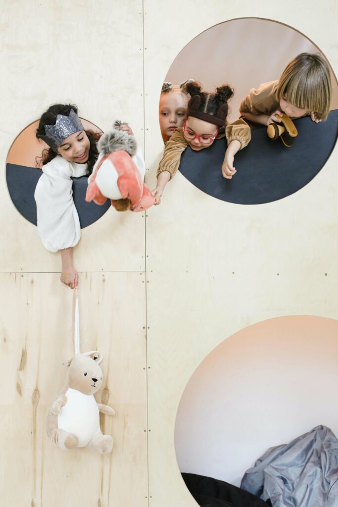 Four children play together on a wooden structure with circular cutouts. One child holds a plush toy, while another dangles a stuffed animal from below, as the others look on and smile.