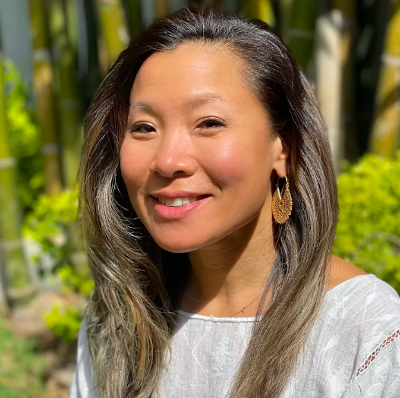 A woman with long, brown hair and gold earrings smiles outdoors. She is wearing a light-colored top, and sunlight highlights her face and hair. Green bamboo and plants are blurred in the background.