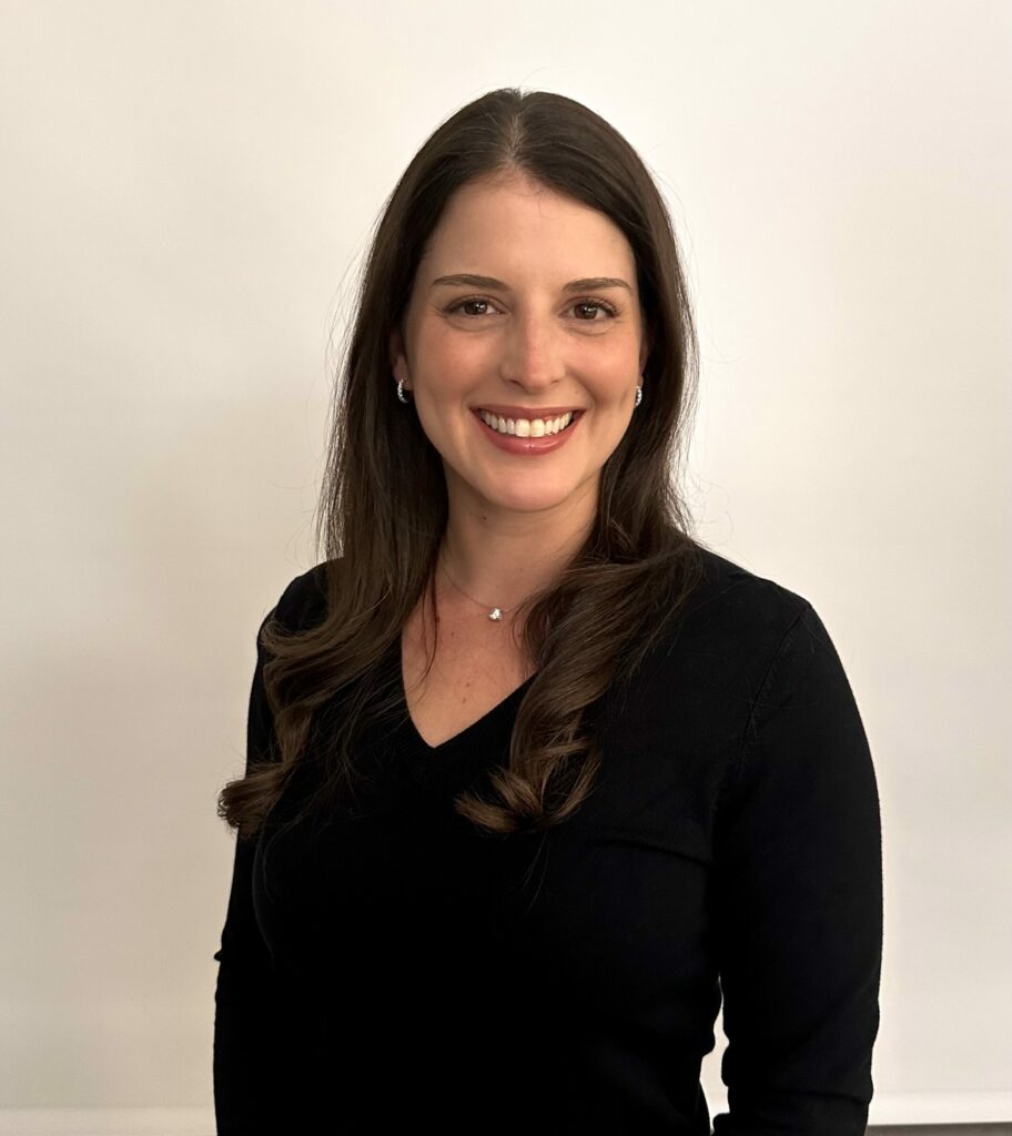 A woman with long brown hair, wearing a black top and a small necklace, is standing and smiling in front of a plain, light-colored background.